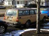 An old, beige volvo station wagon, with a boxy look, in good shape, with red hat linux and apple computer stickers on the rear window, sun reflecting off the rear window, snow on the ground, and a white house in the background