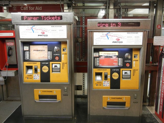 Two electronic PATCO ticket machines, with metal, yellow, and black surfaces and lots of color, a red PATCO station in the background