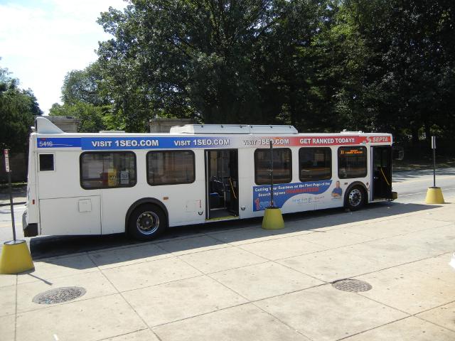 Empty bus with doors open, next to a wide sidewalk terrace with no people, trees in the background