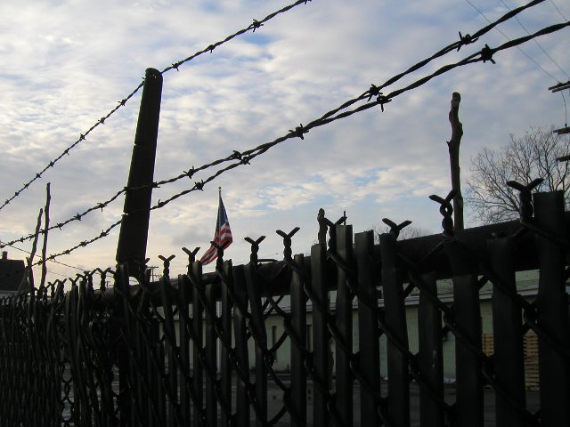 An American flag, flying behind a chain-linked fence with barbed wire, and light clouds against a blue sky