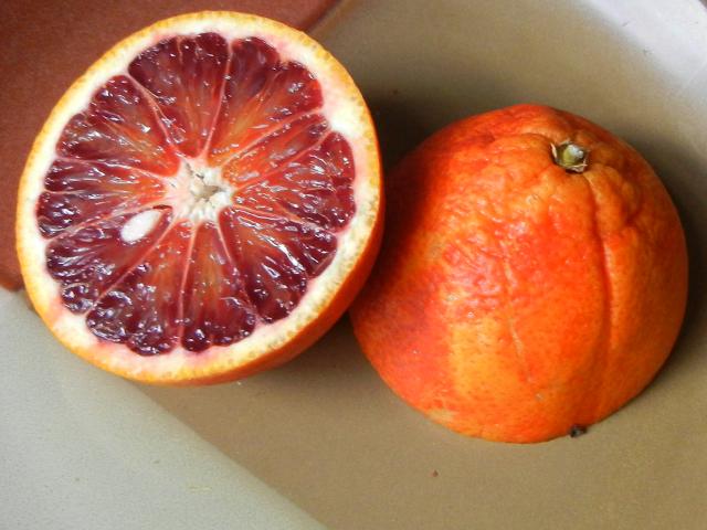 A blood orange sliced in half, showing a cross-section of the interior with a deep red hue, and one sliced seed, and the other half showing the skin with a reddish tinge
