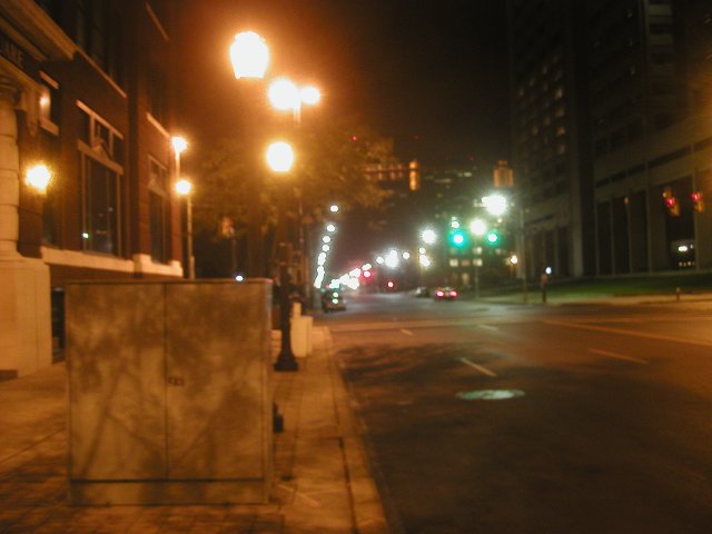 City street at night, lined with streetlights casting a dull orange light, a almost no car traffic, and a red brick building on the left