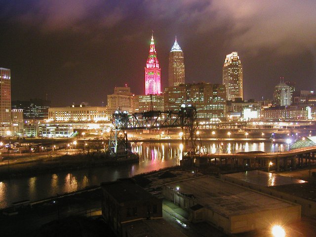 Photo of Cleveland skyline at night, one of the three tallest buildings illuminated bright red with some green, river in foreground, dark drawbridge, and numerous orange-colored lights, reflecting off the river, clouds reflecting a purplish color from the
