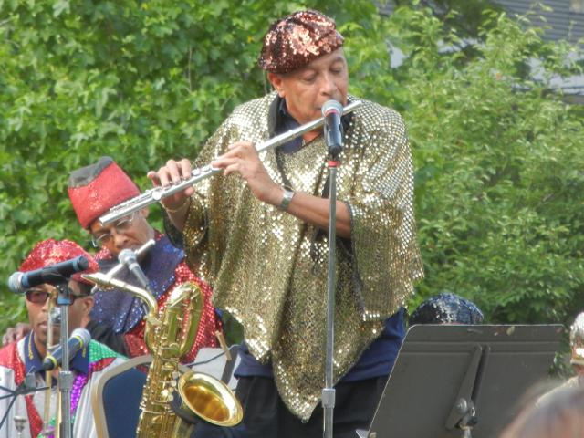 Photo of an older man with a gold sequined robe and a red sequined hat, playing flute in front of a microphone, with two musicians in similar, brightly colored outfits on the left, and trees in the background