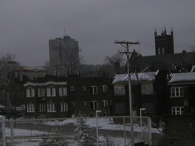 Three two-story apartment buildings, the middle one boarded up, with snow on the ground, and a high-rise building and a church in the background