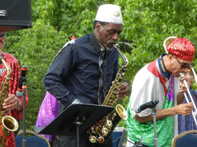An older black man playing alto saxophone, with a white sequined fez, surrounded by a band of musicians in brightly colored, shiny outfits, with a music stand and microphone in the foreground trees in the background