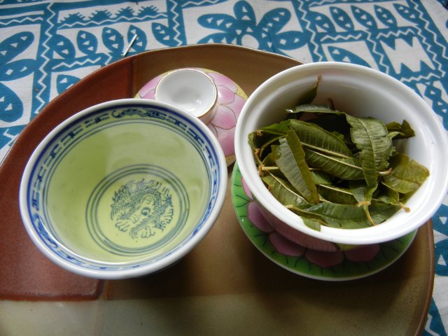 A gaiwan, a Chinese lidded bowl, open, filled with wet lemon verbena leaves, and a cup with a pale greenish-yellow liquid in it, the infusion of the lemon verbena