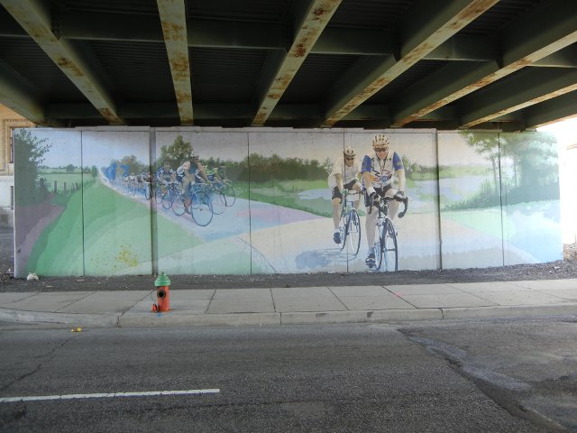 A mural painted on concrete under a bridge, depicting bicyclists in a bicycle race