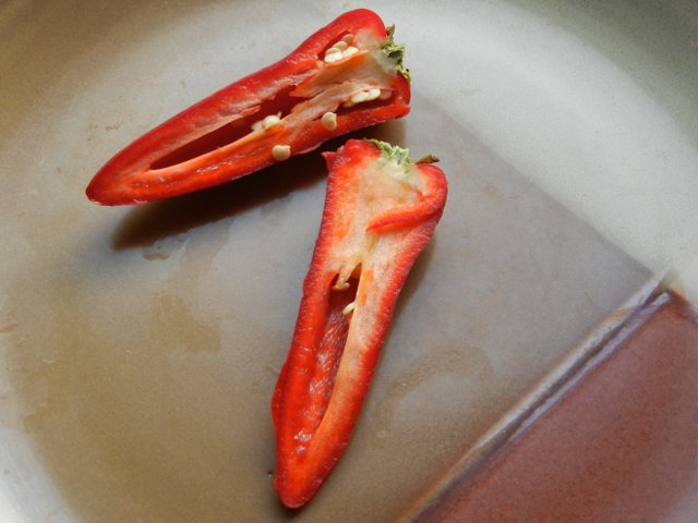 A sliced red fresno pepper, cut in half, showing the pepper interior, intense red color, green stem, straight conical shape, and a few seeds at the top, on a brown ceramic plate