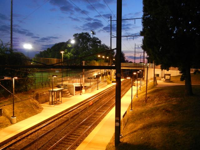 A train station just after dusk, showing tracks and a low platform, with an illuminated athletic field on the left, and a few lingering colors of sunset in the distance