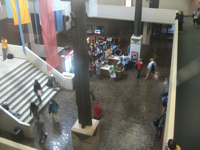 Interior of a college classroom building from the seventies, viewed from two stories above, showing a lobby with large staircase, and some students walking about
