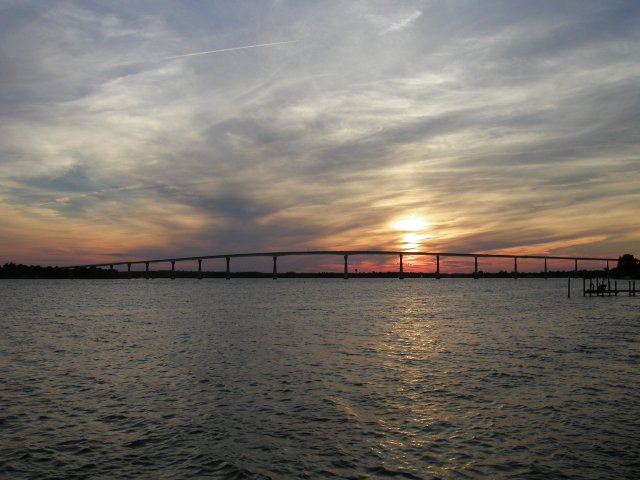 A long bridge over water, with a pink and orange sunset behind clouds, and a gray-blue and white, marbly-looking sky
