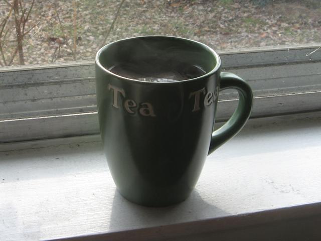 A mug of tea, with an inscription reading Tea, on a windowsill, winter leaves on grass out the window