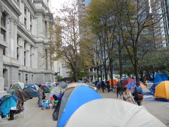 Rows of colorful tents on a plaza, with Philadelphia's city hall on the left, and some mostly bare trees in the background, with tall buildings behind them