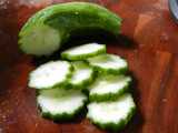 A small cucumber, seedless, with dark green wobbly edge, sliced and slices sitting on a dark wooden cutting board