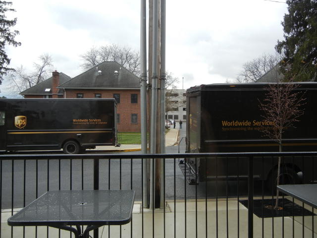 Two UPS trucks, facing opposite directions, parked on opposite sides of a street, with a metal railing in the foreground and a building in the background