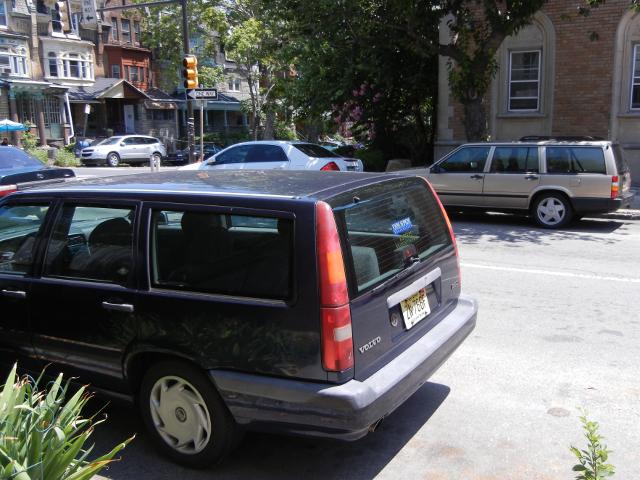 A navy blue volvo station wagon on the near side of a street and a beige volvo station wagon parked opposite, across the street, in an urban residential neighborhood