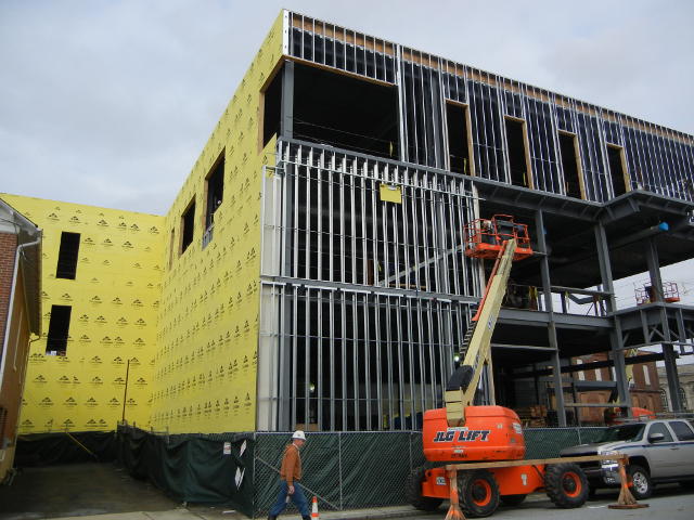 A three-story steel frame building under construction, with exposed steel on the main side and a side showing yellow paper covering two walls, and a bit of machinery and a man carying some equipment