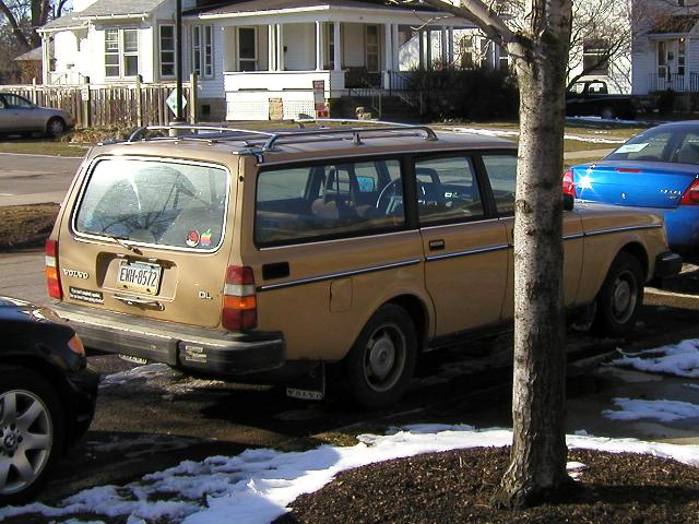An old, beige volvo station wagon, with a boxy look, in good shape, with red hat linux and apple computer stickers on the rear window, sun reflecting off the rear window, snow on the ground, and a white house in the background