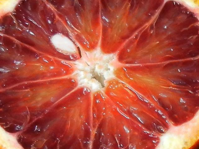 Closeup of a sliced blood orange, showing the center of the orange and the various sections, with a deep reddish tinge to a dark orange color, showing a single seed sliced in half