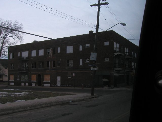 A boarded up three story red brick apartment building with missing windows on the second floor, and balconies overlooking the street, across the street on the left, a vacant lot
