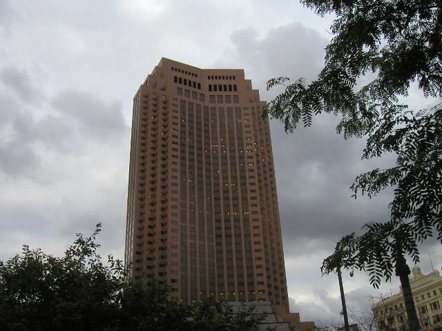 Wide brown skyscraper with black windows, a few yellow lights, against a sky with billowy, dark clouds, with a few leaves of locust foliage around the side of the photo