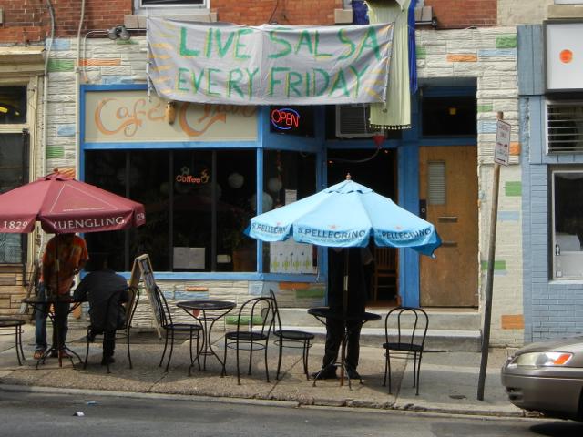 A storefront with some outdoor seating with two umbrellas, a banner hung above the storefront reading: LIVE SALSA EVERY FRIDAY