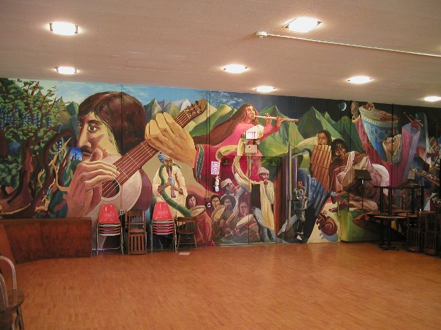 A brightly colored mural depicting musicians from many different cultures, in a room with a plain floor and ceiling and some chairs around the edge
