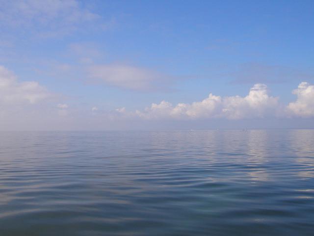 Dark blue open water, with only very tiny ripply waves, and open sky with a few white, billowy clouds, a few tiny boats visible on the horizon