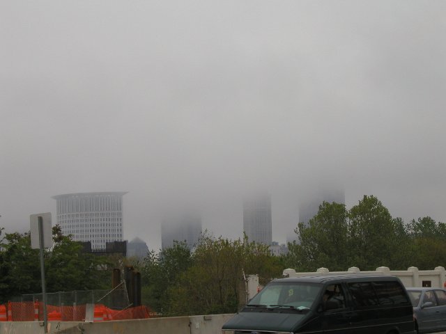 Three tall skyscrapers rising into dense clouds that obscure their tops, one building fully visible, but hazy, on the left, and some trees, a bridge, and construction in the foreground.