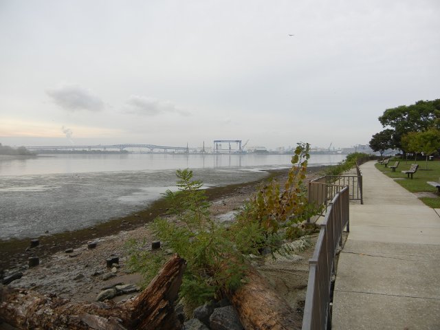 A walkway with a river on the left, a bridge and some industrial stuff in the distance
