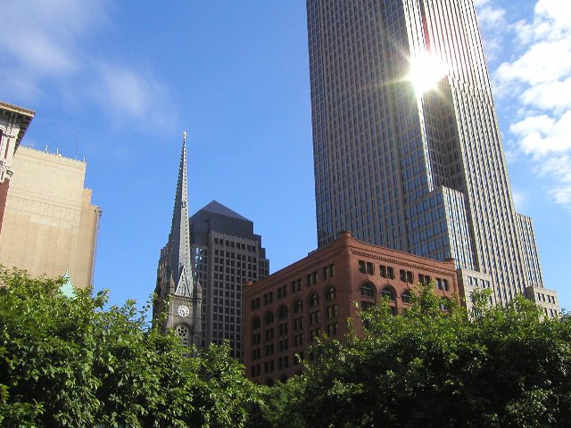 Shiny new skyscraper in back, old red building in front, and an old stone church's steeple, all rising above the tops of green trees