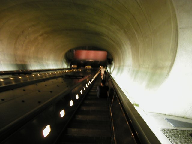 Looking down a very long escalator, with some lights, a round, concrete ceiling above, and a small metal grate on the right