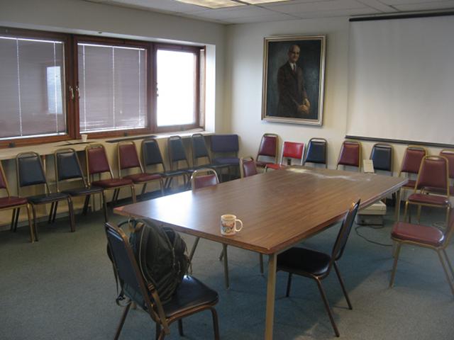 A seminar room at a university, showing a rectangular table with some chairs, and chairs around the edge of the room, a projector screen, and a painting of an older man