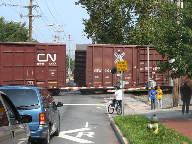 A freight train with boxy, brown cars, with two minivans, a bicyclist, and a couple students all waiting at the gates of the railroad crossing