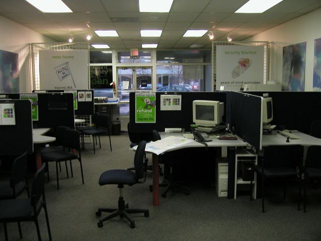 Inside of an H&R Block office, with cubicles with computers, plain  gray carpet, flourescent panel lights in the ceiling, looking out through a glass storefront onto the street in the distance