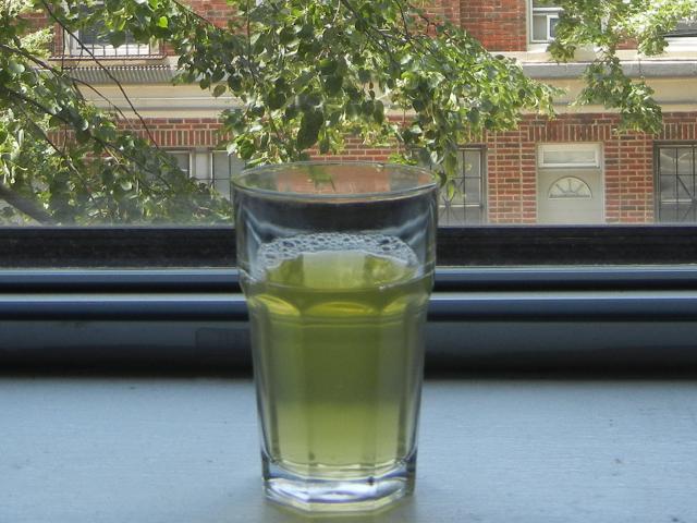 A glass of iced green tea, with no ice, on a windowsill with trees and a brick building in the background