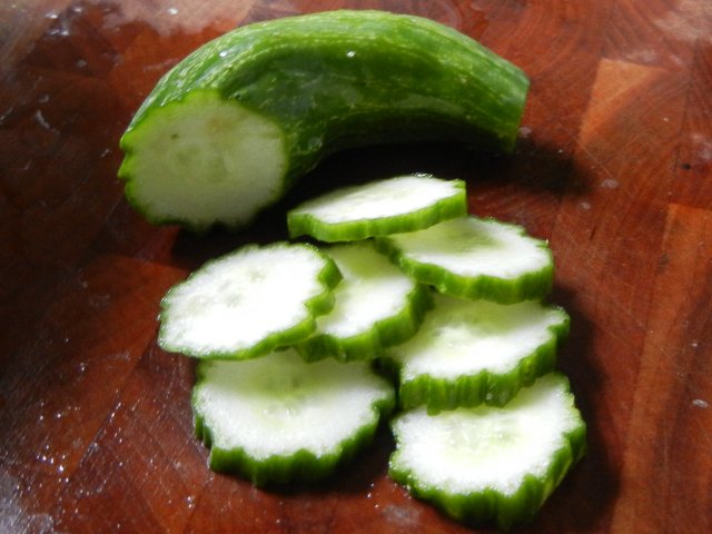 A small cucumber, seedless, with dark green wobbly edge, sliced and slices sitting on a dark wooden cutting board