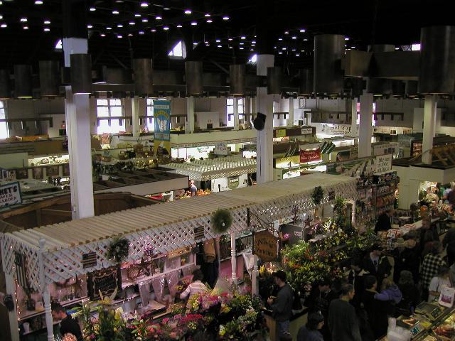 An interior view looking down on a market in a large, open building, showing rows of stands and people in an aisle