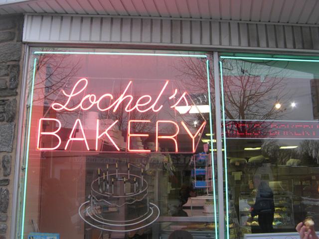 A storefront with a neon sign reading: Lochel's Bakery, with the interior of the bakery faintly visible through the window