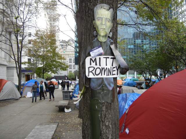 A picture of Frankenstein, labelled Mitt Romney, attached to a tree, surrounded by tents in a city