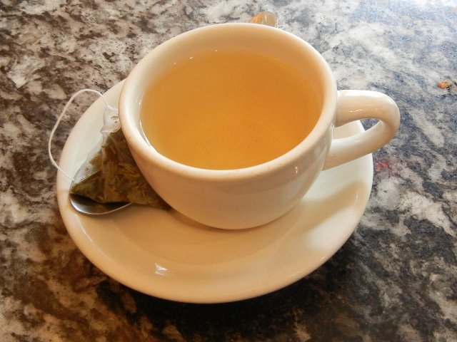A white tea cup with golden-colored tea, on a saucer with a pyramid tea bag and spoon, on a marble surface