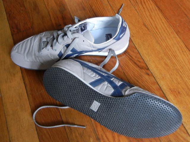 A pair of Onitsuka Tiger (Asics) Serrano Sneakers, gray with navy patterning, a lightweight retro sneaker, one flipped over to show stippled rubber sole for traction, on a wooden floor