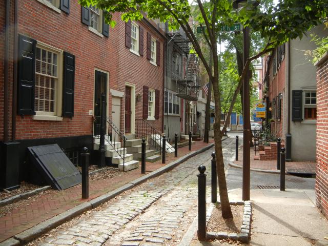 A tiny cobblestone alley with brick streets, with old brick rowhouses, and a paved side-street going to the right