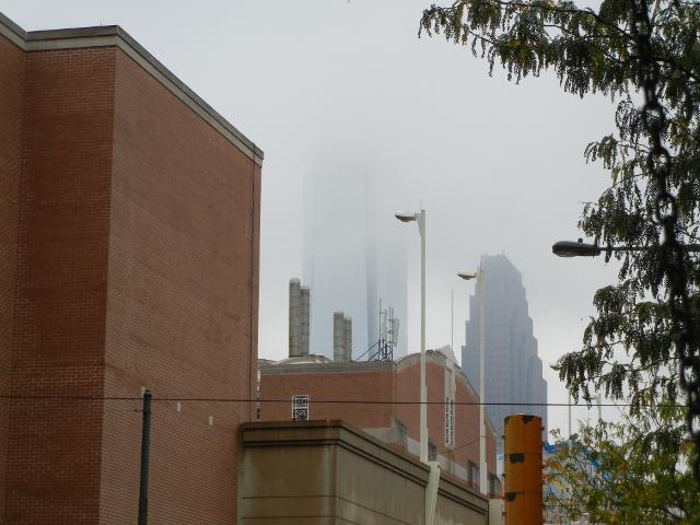 A modern skyscraper rising into a cloud, with its top not visible, a large brick building on the left