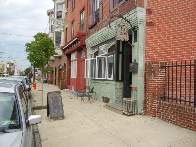 A green-painted storefront with a small sign reading: random tea room, in a row of brick houses, worn down, but ornate, on a city street