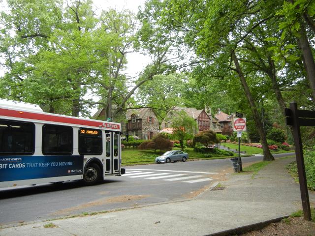 A SEPTA bus in a neighborhood with huge trees and ornate, old houses, approaching a sign that shows both a bus stop and a do-not-enter sign