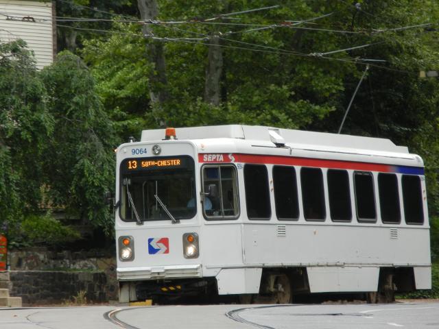 A SEPTA Trolley, white with the red and blue SEPTA logo, showing overhead wires and trees in the background, with the marquee reading: 13 58th-CHESTER