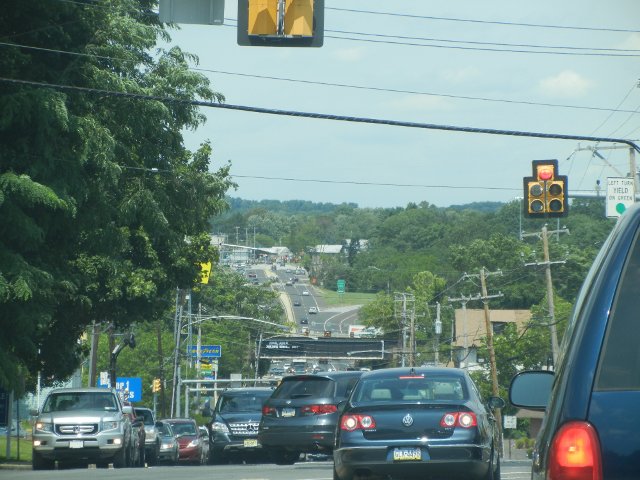 A four-lane road looking down a hill, with tons of cars in both directions, an underpass with a freight train going over it, and trees in the background