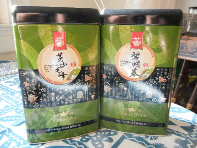 Two 6oz tins of green tea, green and black, one  saying huangshan maofeng and the other Bilouchun (misspelled)